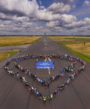 Eine große Gruppe von Menschen steht auf einer Landebahn und bildet mit Fahrrädern ein Herz. In der Mitte des Herzens ist ein Banner mit der Aufschrift „HERZLICH WILLKOMMEN 2026“. Der Himmel ist bewölkt und die Umgebung zeigt grünes Gras.