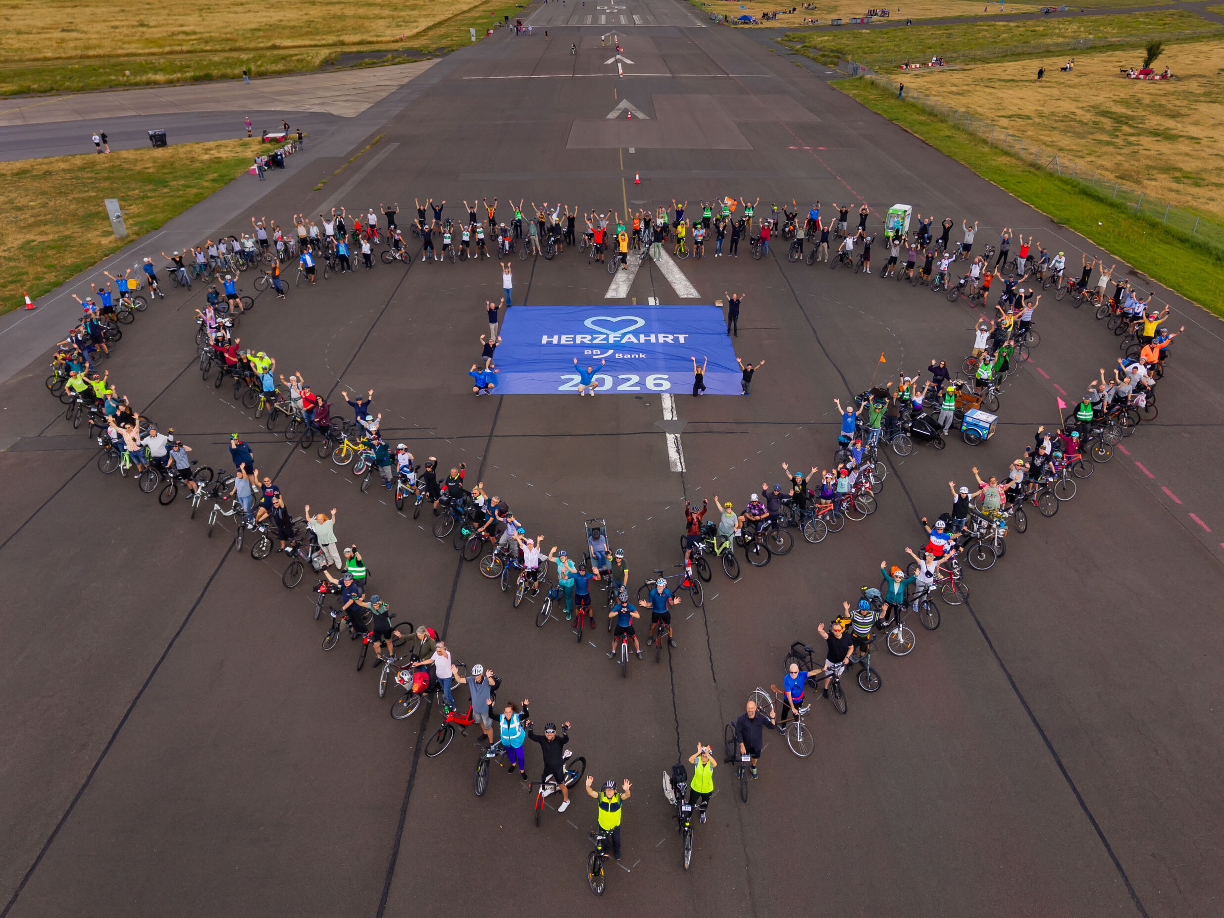 Eine große Gruppe von Menschen steht auf einer Landebahn und bildet mit Fahrrädern ein Herz. In der Mitte des Herzens ist ein Banner mit der Aufschrift „HERZLICH WILLKOMMEN 2026“. Der Himmel ist bewölkt und die Umgebung zeigt grünes Gras.