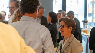 Eine Person in einem grauen Blazer steht in einer Gruppe von Menschen und lächelt. Die Umgebung wirkt lebhaft und gesellig, passend zu einem Networking-Event. Im Hintergrund sind weitere Gesprächspartner zu sehen, die in angeregten Gesprächen vertieft sind.