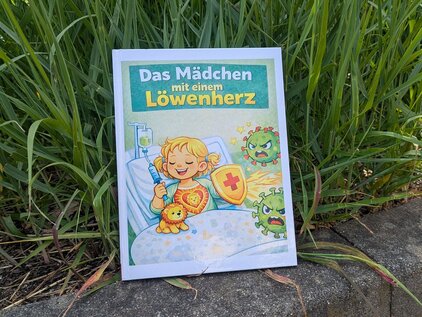 Ein Kinderbuch mit dem Titel „Das Mädchen mit einem Löwenherz“ zeigt ein fröhliches Mädchen im Krankenhaus, das ein Löwenkostüm trägt und ein Kuscheltier umarmt. Um sie herum sind freundliche, cartoonartige Viren abgebildet, was eine positive und mutige Botschaft vermittelt.