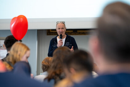 Eine Person hält ein Mikrofon und spricht vor einem Publikum in einem Raum. Im Hintergrund ist ein rotes Herzluftballon sichtbar. Die Zuhörer sind teilweise unscharf dargestellt, was auf die Interaktion zwischen der sprechenden Person und dem Publikum hinweist.
