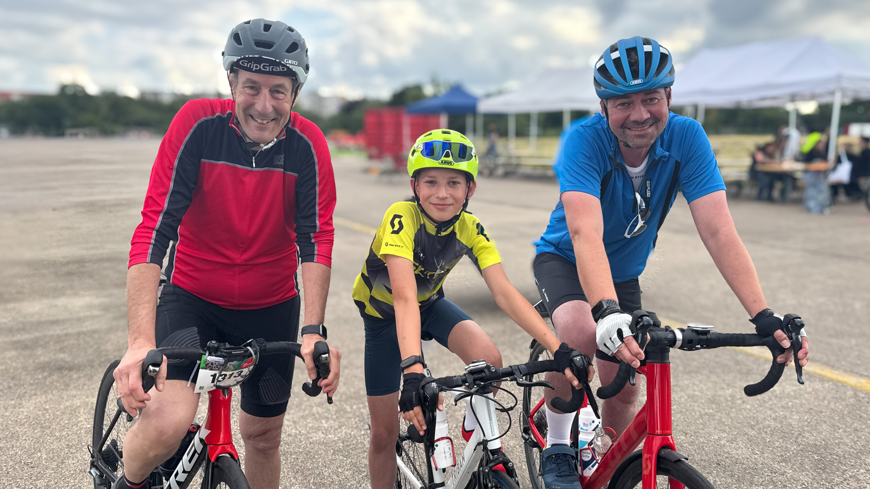 Drei Radfahrer stehen nebeneinander auf einem Parkplatz. Zwei Erwachsene und ein Jugendlicher lächeln in die Kamera. Sie tragen Helme und Sportbekleidung. Der Jugendlicher sitzt auf einem Fahrrad in gelber und schwarzer Kleidung, die Erwachsenen auf eigens für das Radfahren konzipierten Rädern.