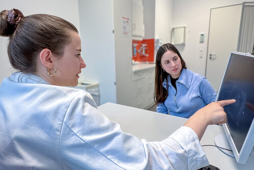Eine Frau zeigt auf einen Computerbildschirm, während eine andere Frau ihr aufmerksam zuhört. Beide sind in einem modernen Büroumfeld, und der Fokus liegt auf dem Gespräch und der Interaktion zwischen den beiden.