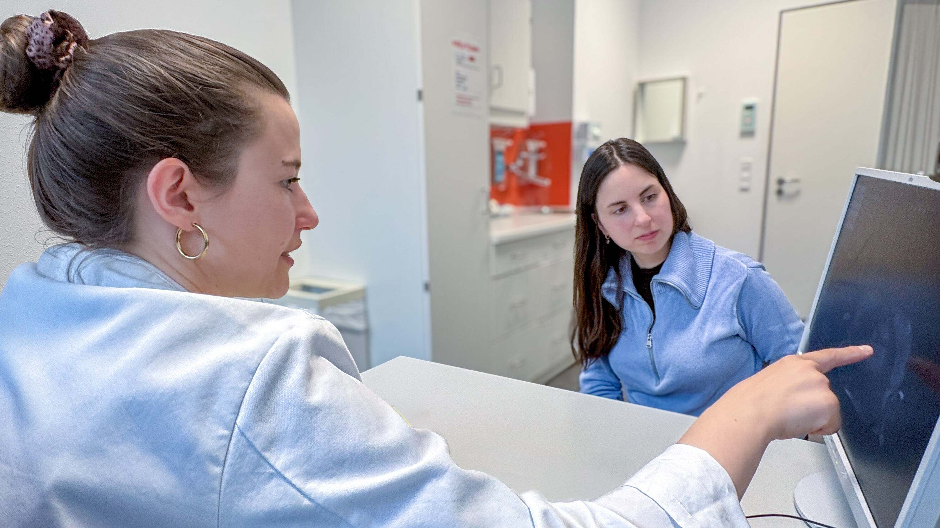 Eine Frau zeigt auf einen Computerbildschirm, während eine andere Frau ihr aufmerksam zuhört. Beide sind in einem modernen Büroumfeld, und der Fokus liegt auf dem Gespräch und der Interaktion zwischen den beiden.