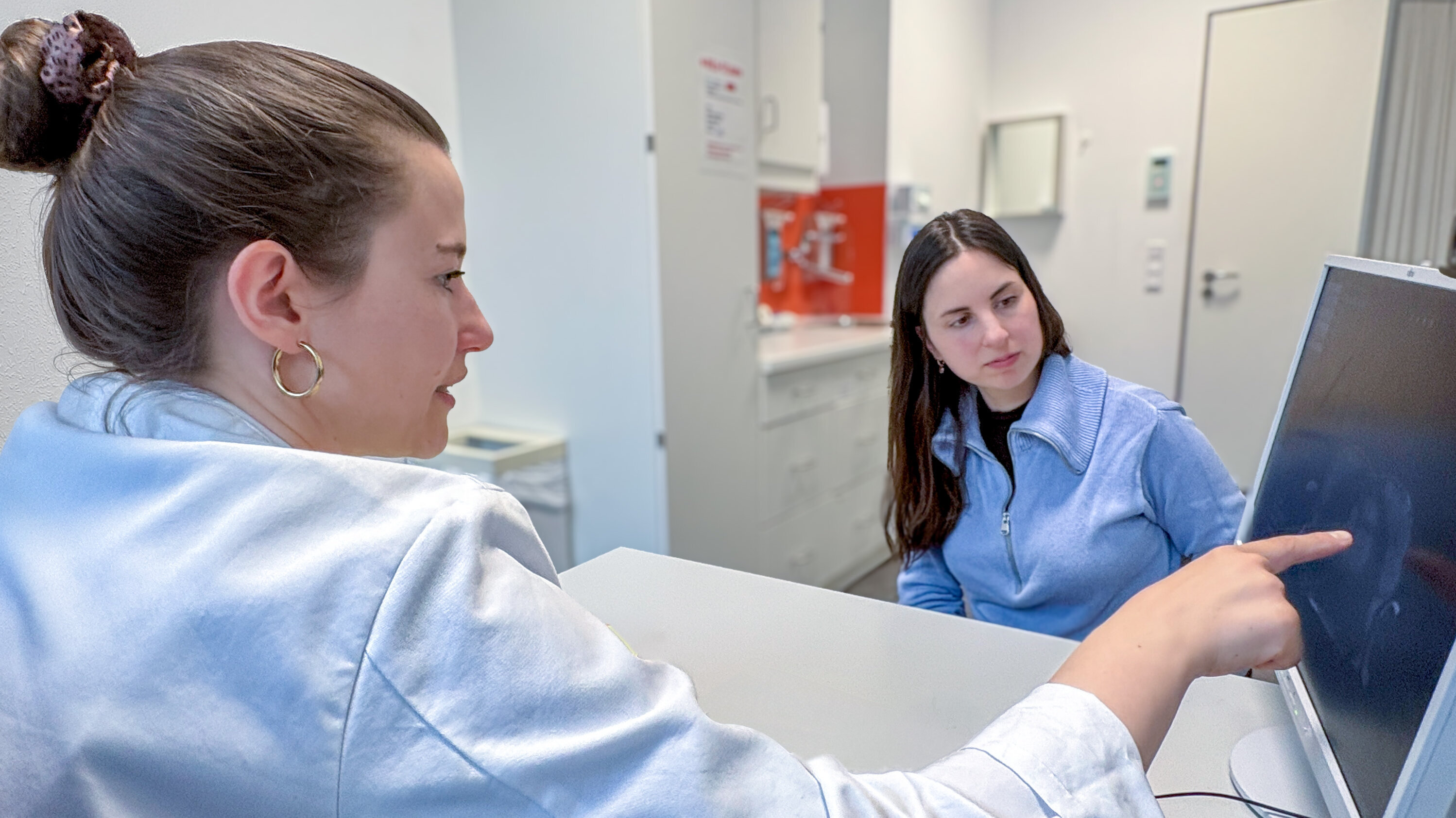 Eine Frau zeigt auf einen Computerbildschirm, während eine andere Frau ihr aufmerksam zuhört. Beide sind in einem modernen Büroumfeld, und der Fokus liegt auf dem Gespräch und der Interaktion zwischen den beiden.