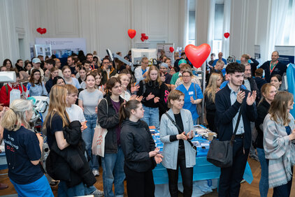 Eine Menschenmenge steht in einem Raum, applaudiert und interagiert miteinander. Einige Personen tragen T-Shirts mit Logos. Im Hintergrund sind Informationsstände und Dekorationen mit Herzballons zu sehen. Die Atmosphäre wirkt lebhaft und engagiert.