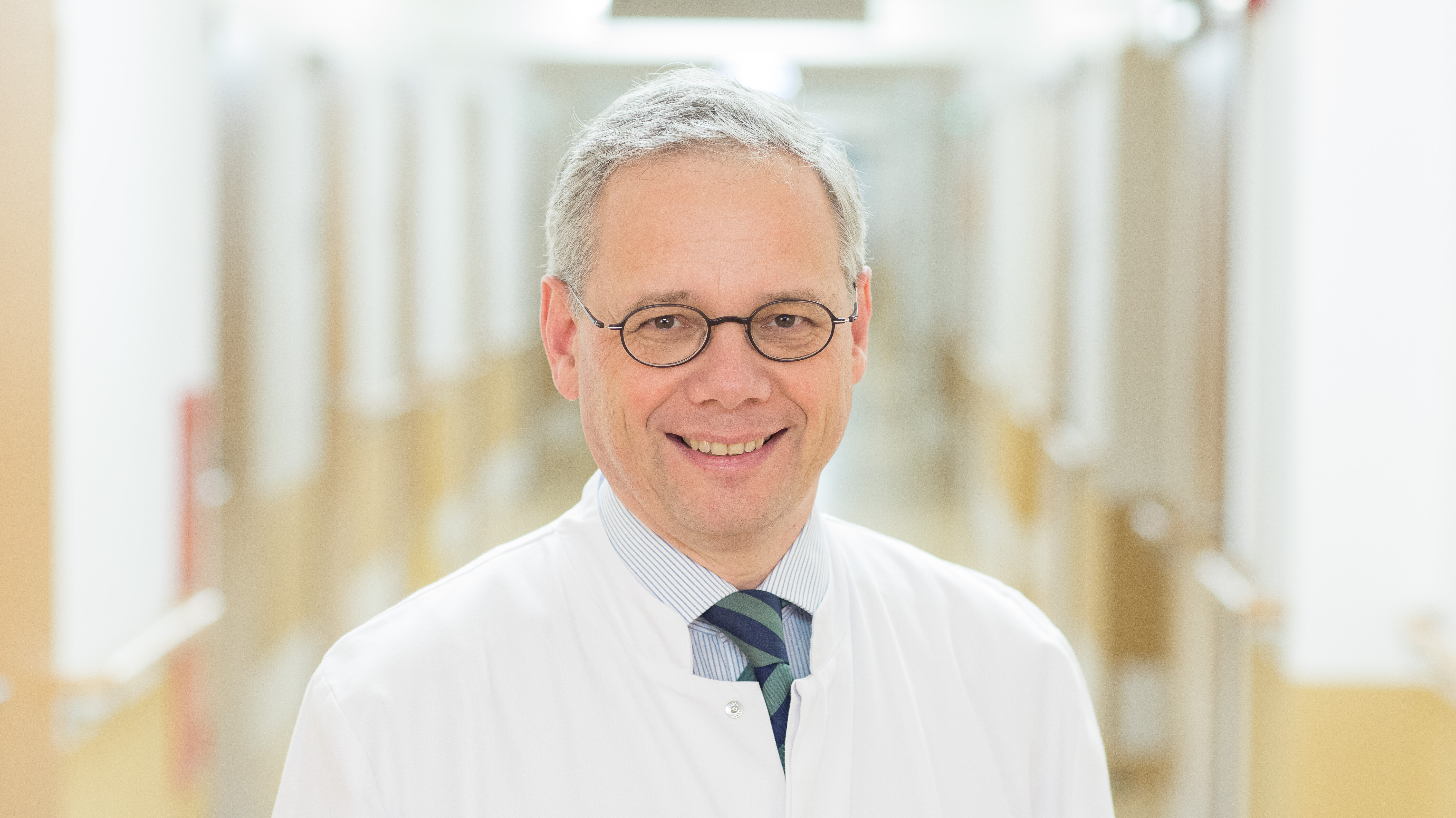 Ein Arzt lächelt in einem Krankenhausflur. Er trägt einen weißen Kittel und eine Krawatte. Der Hintergrund zeigt verschwommen die Wände des Flures, was auf eine medizinische Umgebung hinweist.