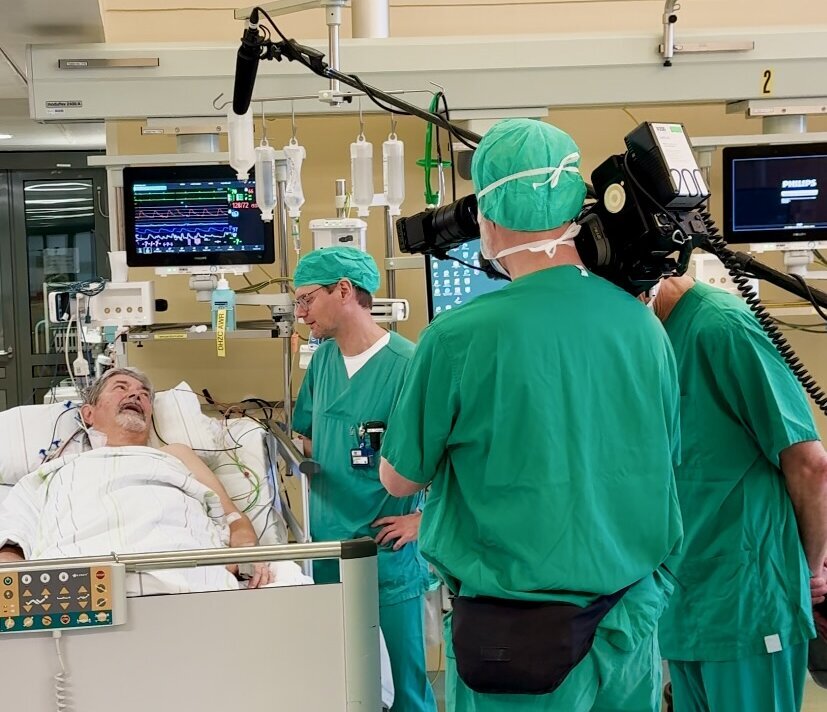 Im Bild ist ein Patient im Krankenhausbett zu sehen, umgeben von Ärzten in grünen Kitteln. Ein Arzt spricht mit dem Patienten, während ein anderer einen Kameramann beobachtet, der die Szene filmt. Monitore zeigen medizinische Daten im Hintergrund.