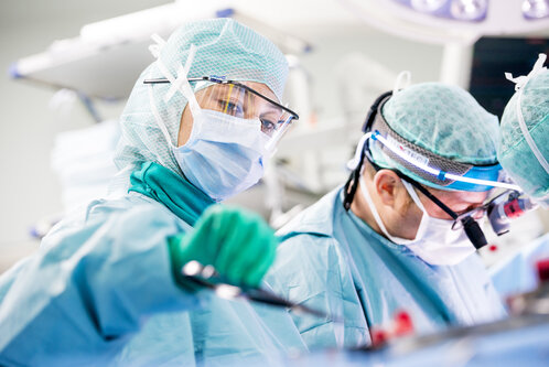 Zwei Chirurgen in Schutzkleidung führen eine Operation durch. Der Fokus liegt auf einem Chirurgen, der ein medizinisches Instrument hält, während der andere intensiv auf die Operation schaut. Beide tragen Masken und Schutzbrillen in einem modernen OP-Saal.