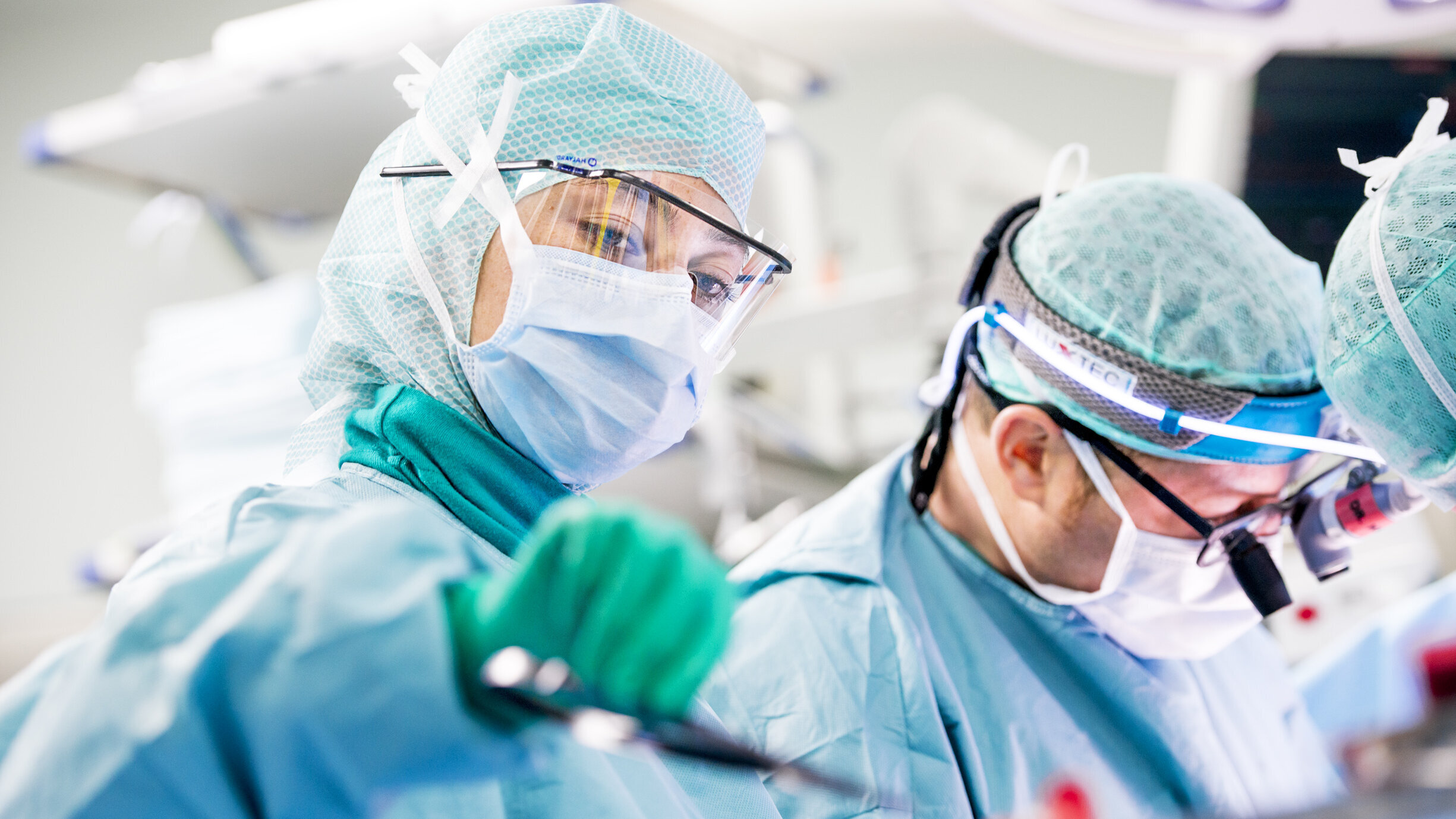 Zwei Chirurgen in Schutzkleidung führen eine Operation durch. Der Fokus liegt auf einem Chirurgen, der ein medizinisches Instrument hält, während der andere intensiv auf die Operation schaut. Beide tragen Masken und Schutzbrillen in einem modernen OP-Saal.