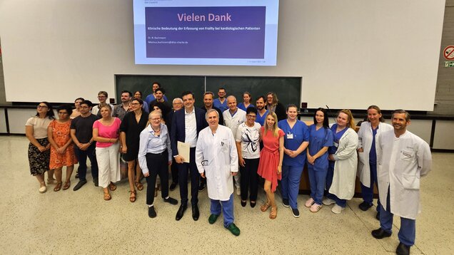 Auf dem Bild stehen mehrere Personen in einem Seminarraum zusammen, einige in medizinischer Kleidung. Im Hintergrund ist eine Präsentation mit dem Titel „Vielen Dank – Klinische Bedeutung der Erfassung von Frailty bei kardiologischen Patienten“ zu sehen. Die Gruppe wirkt freundlich und engagiert.