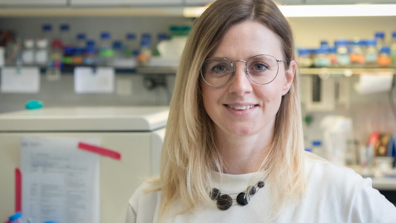 Eine lächelnde Frau mit langen, blonden Haaren steht im Labor. Sie trägt eine helle Bluse und eine auffällige Halskette. Im Hintergrund sind Laborausrüstung und Reagenzgläser sichtbar, die auf eine wissenschaftliche Umgebung hinweisen.