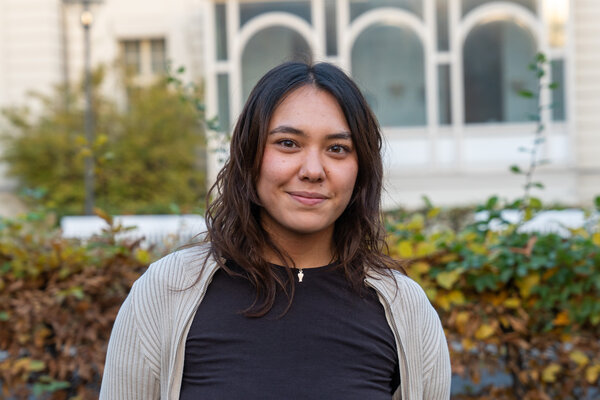 Eine junge Frau mit langen, lockigen Haaren lächelt in die Kamera. Sie trägt ein schwarzes T-Shirt und einen hellen Pullover. Im Hintergrund sind einige Pflanzen und ein Gebäude sichtbar.