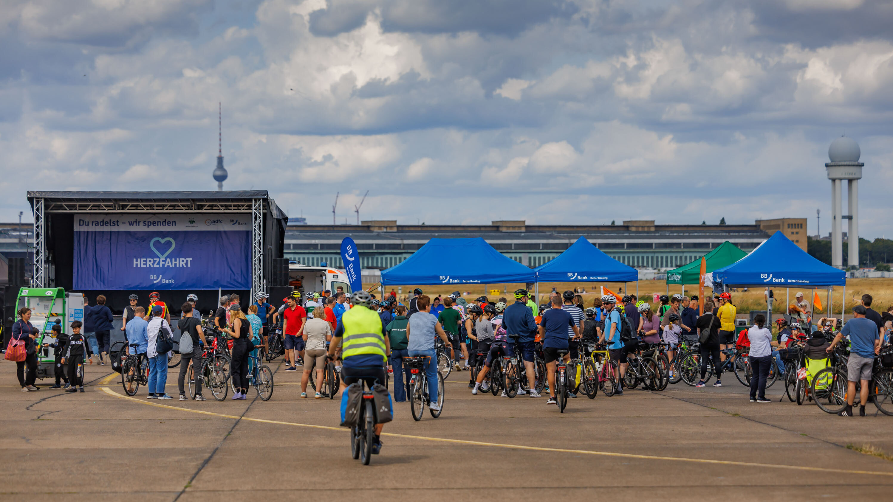 Eine große Gruppe von Menschen steht mit Fahrrädern auf einem Platz, während vor ihnen eine Bühne aufgebaut ist. Im Hintergrund sind Zelte und ein bekanntes Berliner Gebäude zu sehen. Das Event scheint eine Fahrradtour oder eine Versammlung zu sein, möglicherweise zum Thema Gesundheit.