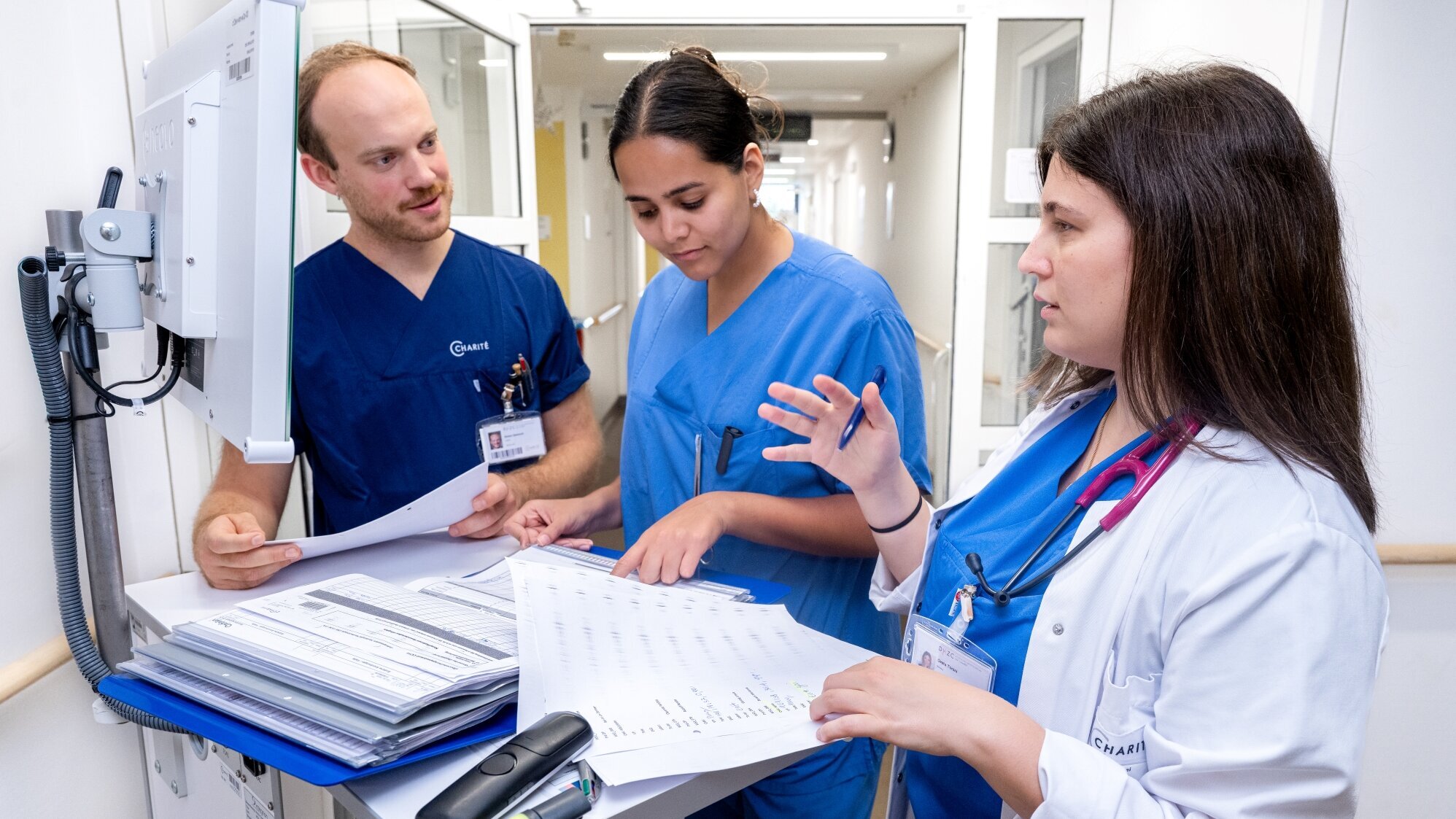 Drei medizinische Fachkräfte in blauen Uniformen stehen an einem Informationsschalter. Sie besprechen Unterlagen und Notizen, während sie sich auf einen Bildschirm im Hintergrund konzentrieren. Einige Dokumente sind auf dem Tisch verteilt, und ein Stethoskop ist sichtbar.