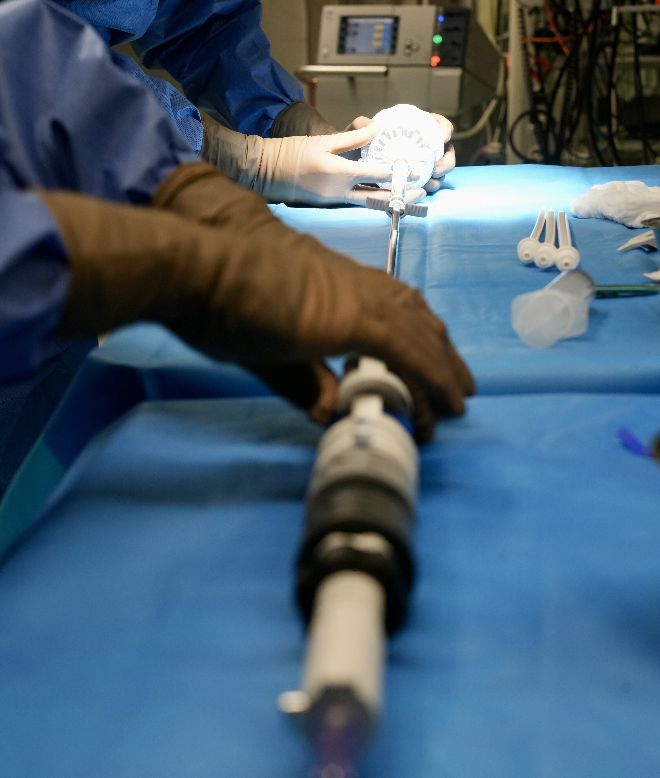 Zwei Hände, die in chirurgischen Handschuhen und blauen Kitteln gekleidet sind, arbeiten an einem medizinischen Gerät auf einem Operationsstuhl. Der Tisch ist mit einem blauen Tuch bedeckt. Im Hintergrund sind verschiedene medizinische Geräte zu sehen.