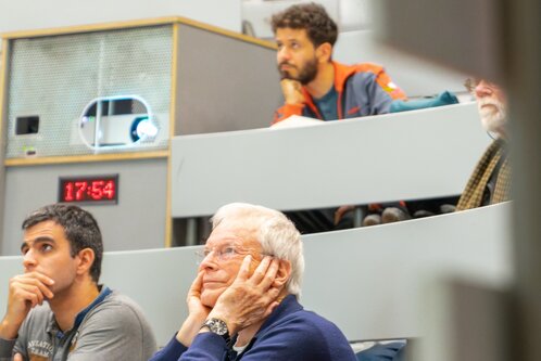 In einem Hörsaal schauen mehrere Personen auf einen Präsentationsbildschirm. Ein älterer Mann mit grauen Haaren stützt seinen Kopf in die Hände, während jüngere Teilnehmer aufmerksam zuhören. Auf einem Tisch ist eine digitale Uhr abgebildet, die auf 17:54 steht.