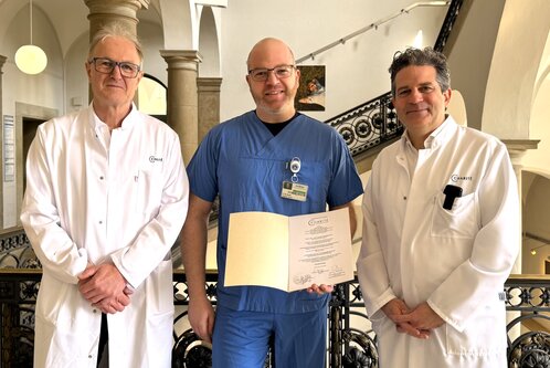 Drei Ärzte stehen in einem Flur zusammen. Der Arzt in der Mitte trägt blaue Arbeitskleidung und hält ein Zertifikat oder eine Urkunde. Die beiden anderen Ärzte tragen weiße Kittel. Alle drei blicken freundlich in die Kamera.