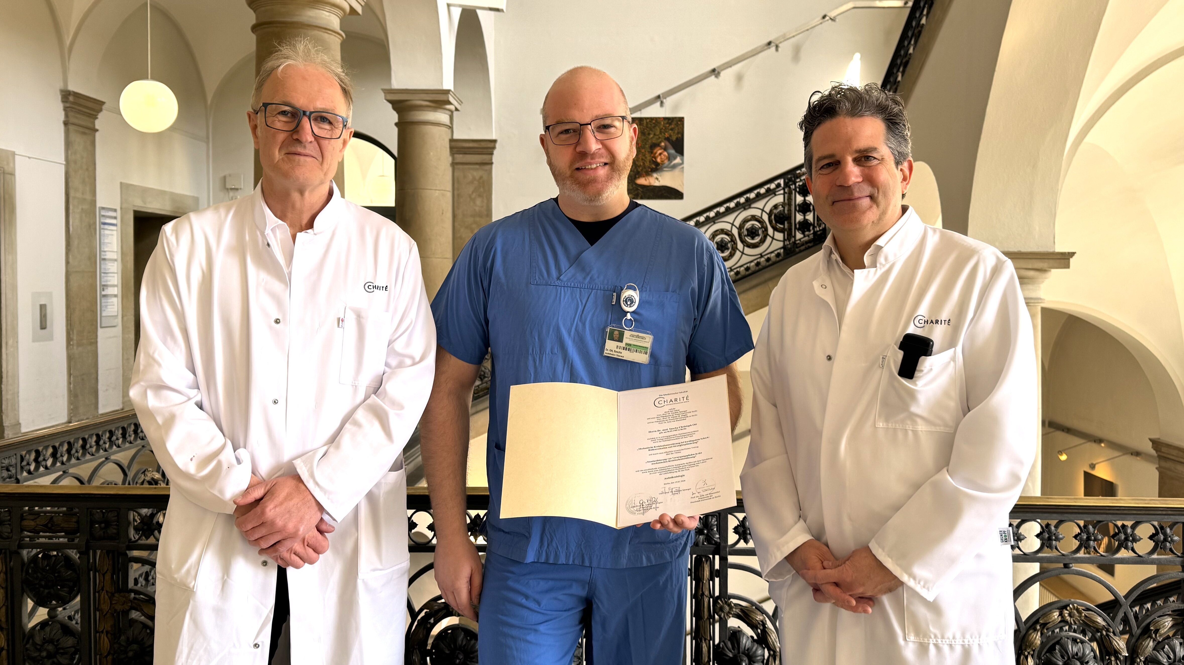 Drei Ärzte stehen in einem Flur zusammen. Der Arzt in der Mitte trägt blaue Arbeitskleidung und hält ein Zertifikat oder eine Urkunde. Die beiden anderen Ärzte tragen weiße Kittel. Alle drei blicken freundlich in die Kamera.