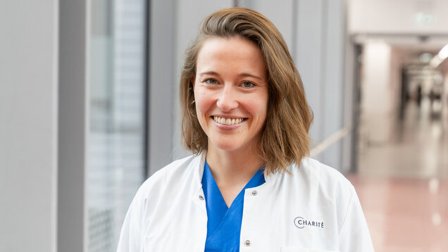 Eine lächelnde Medizinerin trägt einen weißen Kittel über blauer Kleidung. Sie steht in einem modernen Flur mit großen Fenstern im Hintergrund. Ihr freundlicher Gesichtsausdruck vermittelt Offenheit und Vertrauen.