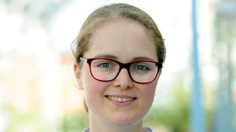 Eine lächelnde Frau in einem weißen Arztkittel steht in einem hellen, unscharfen Outdoor-Hintergrund. Sie trägt eine Brille und hat mittellange Haare. Ihr freundlicher Ausdruck vermittelt Professionalität und Zugänglichkeit.
