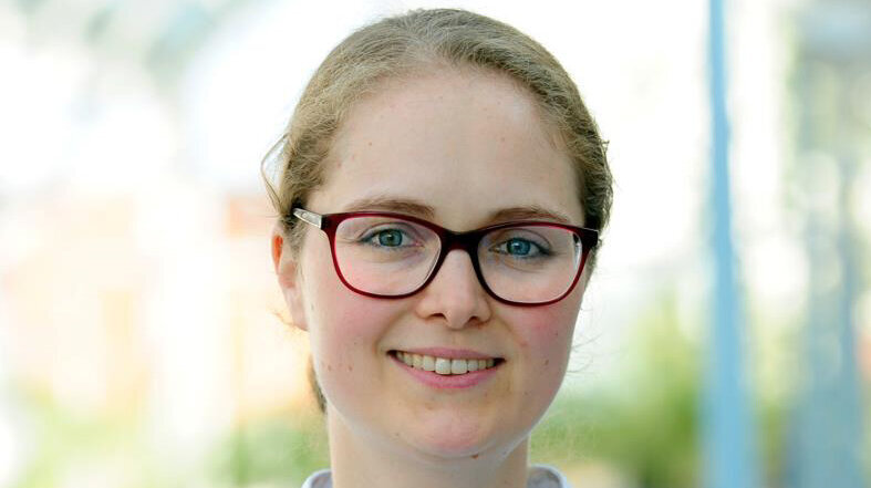 Eine lächelnde Frau in einem weißen Arztkittel steht in einem hellen, unscharfen Outdoor-Hintergrund. Sie trägt eine Brille und hat mittellange Haare. Ihr freundlicher Ausdruck vermittelt Professionalität und Zugänglichkeit.
