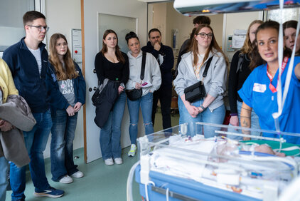 Eine Gruppe von Menschen, darunter Jugendliche und Erwachsene, steht in einem Krankenhauszimmer und hört einer Krankenschwester zu, die einen Informationsvortrag hält. Im Vordergrund befindet sich ein Inkubator mit medizinischen Geräten. Die Atmosphäre ist konzentriert und interessiert.