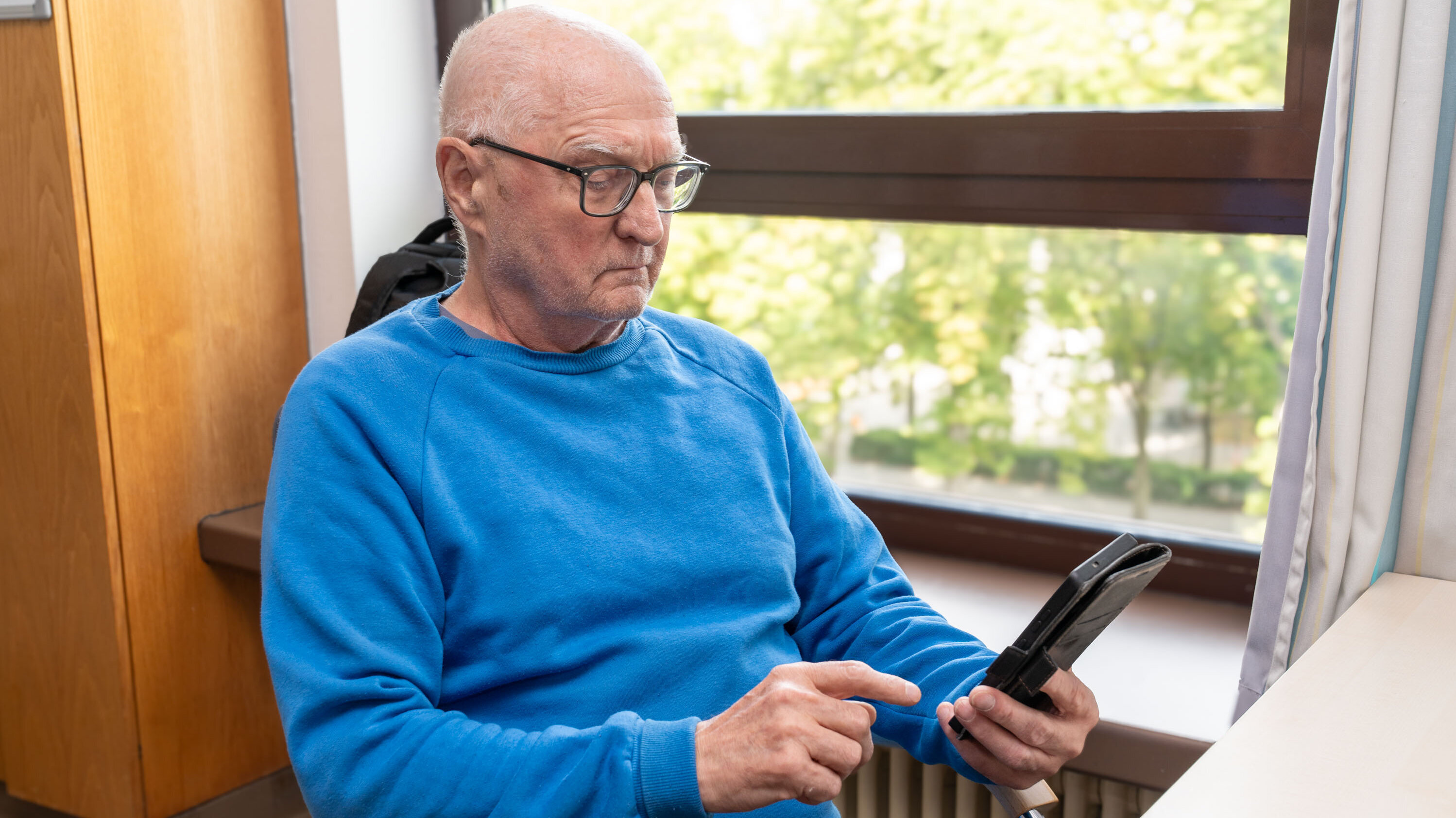 Ein älterer Mann mit Brille sitzt an einem Tisch und schaut konzentriert auf ein Smartphone in seiner Hand. Er trägt einen blauen Pullover und befindet sich in einem hellen Raum mit einem Fenster, durch das grünes Licht strömt.
