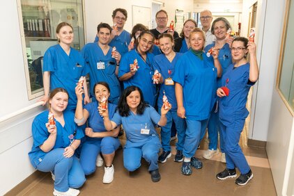 Eine Gruppe von 14 Personen in blauen Krankenhausuniformen steht in einem Flur. Sie lächeln und halten Schokoladenfiguren in der Hand. Einige Personen knien, während andere stehen. Alle wirken freundlich und fröhlich.