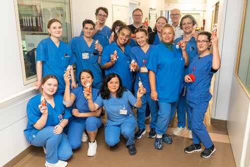 Eine Gruppe von 14 Personen in blauen Krankenhausuniformen steht in einem Flur. Sie lächeln und halten Schokoladenfiguren in der Hand. Einige Personen knien, während andere stehen. Alle wirken freundlich und fröhlich.