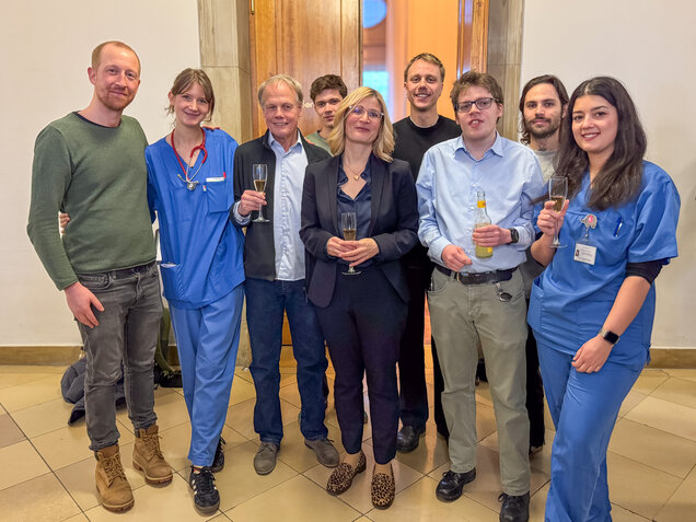 Eine Gruppe von neun Personen steht zusammen und lächelt. Einige tragen Arbeitskleidung im medizinischen Bereich. Sie halten Gläser mit Getränken in der Hand und scheinen einen feierlichen Anlass zu genießen. Der Hintergrund zeigt eine hell erleuchtete Wand.