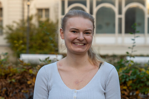 Eine lächelnde Frau mit blonden Haaren, die in einem hellen, eng anliegenden Oberteil gekleidet ist, steht im Freien. Im Hintergrund sind leicht verschwommene Pflanzen und ein Gebäude zu sehen. Die Umgebung wirkt herbstlich mit buntem Laub.