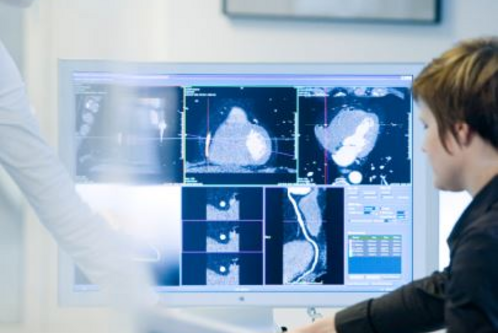 Eine Person sitzt vor einem Computerbildschirm, auf dem mehrere medizinische Bildgebungen wie CT-Scans zu sehen sind. Eine zweite Person zeigt auf den Bildschirm. Die Szene vermittelt den Austausch von Informationen im medizinischen Kontext.