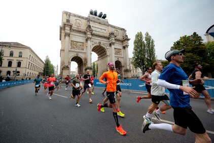 Eine Gruppe von Läufern nimmt an einem Marathon teil und läuft unter einem großen historischen Tor. Im Hintergrund sind Bäume und Gebäude zu sehen, während die Läufer in bunten Sportoutfits unterwegs sind. Die Atmosphäre ist sportlich und dynamisch.