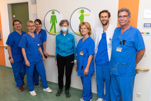 Eine Gruppe von acht Personen steht in einem Krankenhausflur. Die meisten tragen medizinische Kleidung in Blau, während eine Frau in der Mitte eine Maske trägt und Sportbekleidung anhat. Hinter ihnen sind Symbole für Männer und Frauen auf der Wand zu sehen.