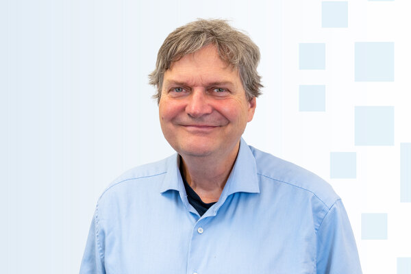 Ein lächelnder Mann mit schulterlangen, grauen Haaren trägt ein hellblaues Hemd. Er steht vor einem sanften, hellen Hintergrund mit geometrischen Formen. Der Mann strahlt Freundlichkeit aus.