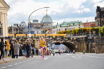 Ein Läufer in gelber Sportkleidung sprintet eine Straße entlang, während Zuschauer auf beiden Seiten stehen und anfeuern. Im Hintergrund sind historische Gebäude und eine Brücke zu sehen, die zur Stadtansicht beitragen. Der Himmel ist teilweise bewölkt.
