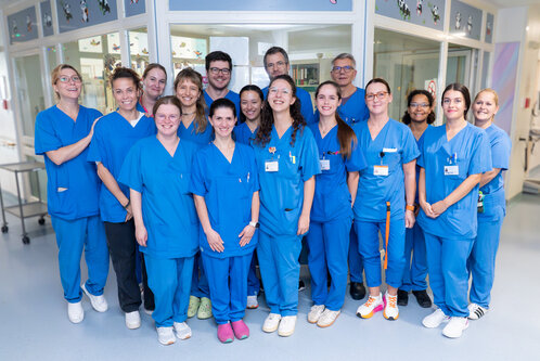 Eine Gruppe von 16 Personen in blauer Arbeitskleidung steht in einem Krankenhaus. Sie lächeln und stehen dicht zusammen, während sie vor einer Wand mit Fenstern posieren. Die Mehrheit der Personen ist weiblich, mehrere tragen Namensschilder.