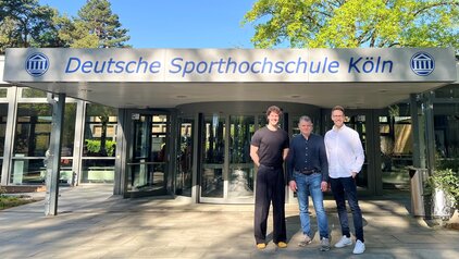 Drei Personen stehen vor dem Eingang der Deutschen Sporthochschule Köln. Über ihnen ist das Schild mit dem Namen der Institution gut sichtbar. Im Hintergrund sind Bäume und eine moderne Gebäudestruktur zu erkennen.