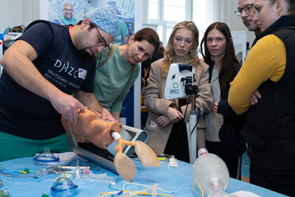 Eine Person demonstriert medizinische Techniken an einem Übungsmaterial in einer Lernumgebung. Eine Gruppe von fünf Personen schaut aufmerksam zu und beobachtet den Prozess. Auf dem Tisch sind verschiedene medizinische Geräte und Utensilien sichtbar.