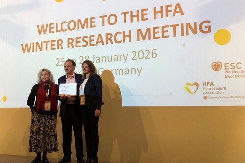 Drei Personen stehen auf einer Bühne vor einem Banner mit dem Titel „Willkommen zum HFA Winter Research Meeting“ und den Daten 26. bis 28. Januar 2026 in Deutschland. Zwei der Personen halten eine Auszeichnung oder ein Dokument in den Händen.