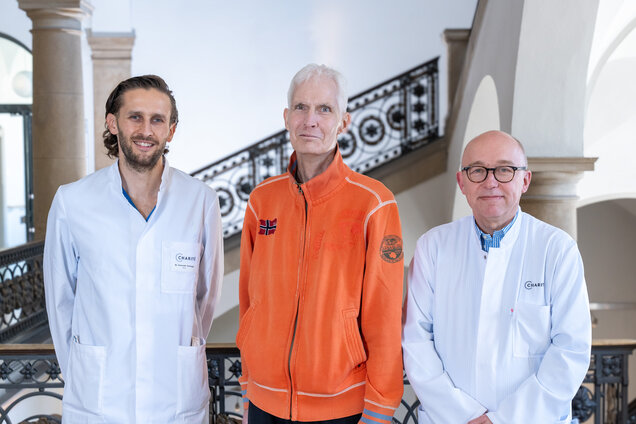 Drei Männer stehen in einem fließenden Raum. Zwei tragen weiße Arztmäntel, der dritte trägt eine orange Jacke. Sie lächeln und stehen nebeneinander, während ein elegantes Treppenhaus im Hintergrund sichtbar ist.