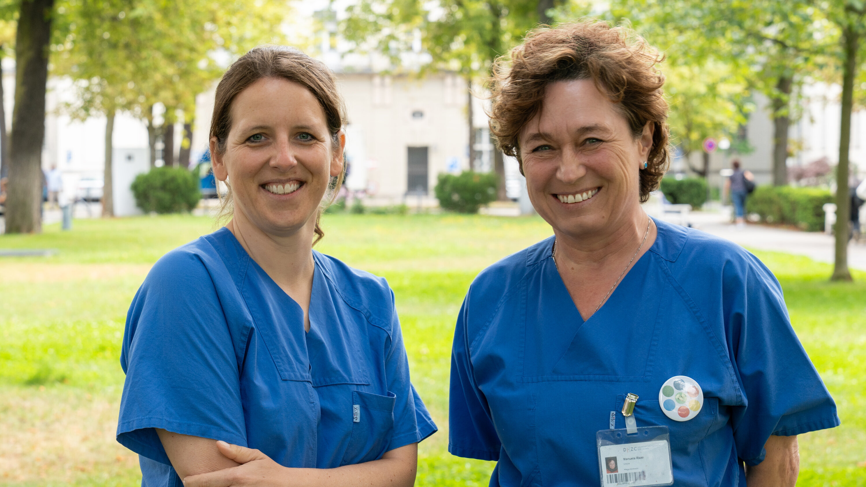 Zwei Frauen in blauen Krankenschwesternuniformen stehen in einem Park. Sie lächeln freundlich in die Kamera. Der Hintergrund zeigt Bäume und grünes Gras, was eine ruhige und einladende Atmosphäre schafft.