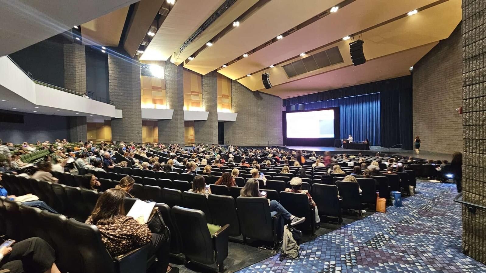 Ein großer Veranstaltungsraum mit vielen sitzenden Personen. Auf der Bühne ist ein Bildschirm mit Präsentationsinhalten zu sehen. Die Zuschauer scheinen aufmerksam zu sein und einige nutzen elektronische Geräte. Die Atmosphäre wirkt konzentriert und informativ.
