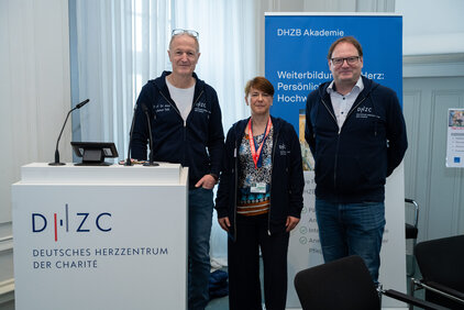 Drei Personen stehen vor einem Präsentationsstand des Deutschen Herzzentrums der Charité (DHZB). Sie tragen schwarze Sweatshirts mit dem DHZB-Logo. Im Hintergrund ist ein Banner mit Informationen zur DHZB Akademie und Weiterbildung zu sehen. 
