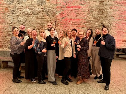 Eine Gruppe von etwa 15 Personen steht zusammen in einem festlichen Raum mit Steinwänden. Alle halten Drinks in den Händen, lächeln und posieren für das Foto. Die Atmosphäre ist fröhlich und feierlich.