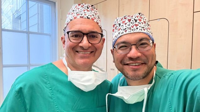 Zwei Ärzte in grünen OP-Kitteln und bunten Haarnetzen lächeln in die Kamera. Sie stehen in einem hellen Raum mit Holzregalen. Beide tragen Mundschutz und zeigen eine freundliche, kollegiale Atmosphäre.