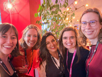 Sechs Frauen stehen gemeinsam in einem festlichen Ambiente, lächeln und posieren für das Foto. Einige halten Weingläser, während eine Pflanze im Hintergrund sichtbar ist. Alle tragen Namensschilder und sind in farbenfroher Kleidung gekleidet.