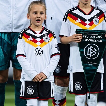 Zwei junge Mädchen stehen in Fußballtrikots der deutschen Nationalmannschaft auf dem Spielfeld. Eines hält ein Wimpel mit der Aufschrift "Deutschland gegen Griechenland". Beide tragen farbige Socken und betonen die Teamfarben. Sie wirken aufgeregt und konzentriert.