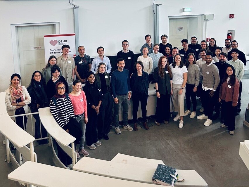 Eine Gruppe von Teilnehmern steht in einem modernen Konferenzraum vor einer Präsentationswand mit der Aufschrift "Young-DZHK Cardiovascular Research Summit 2026, Berlin, 25.09.2026". Die Anwesenden sind vielfältig und tragen unterschiedliche Kleidung, während sie lächeln und in die Kamera schauen.